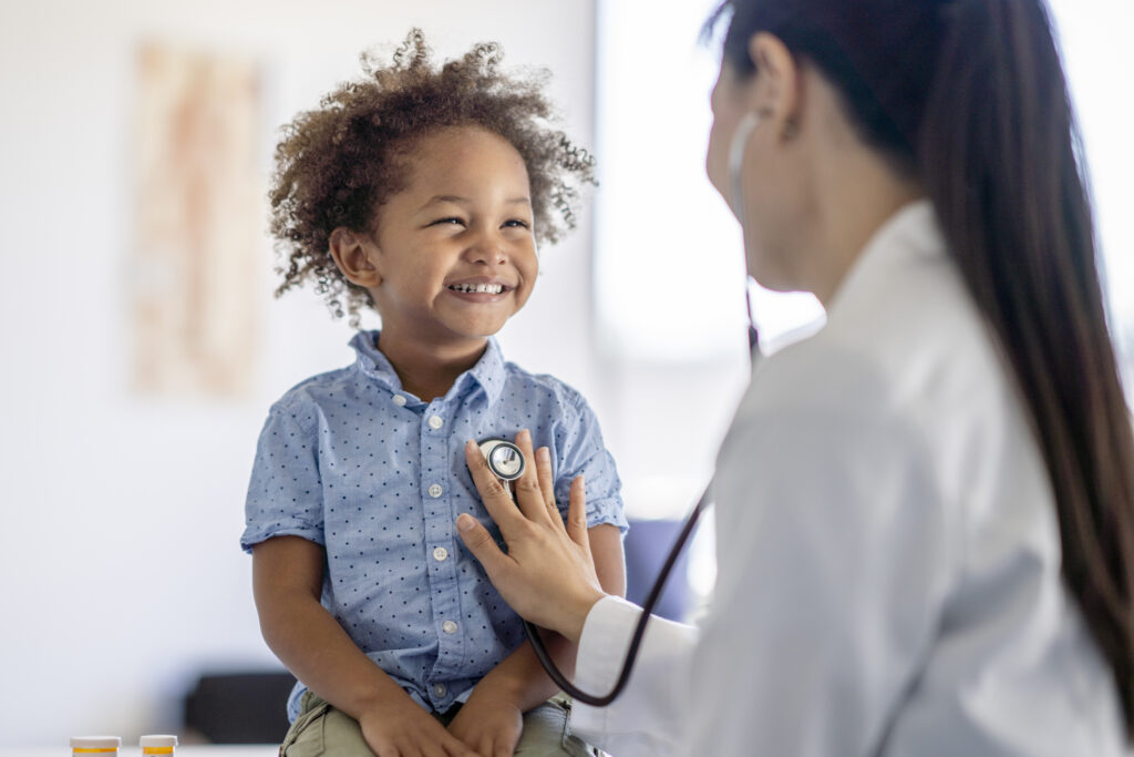 A female doctor of Asian decent listens to her patients heart with her stethoscope. The little boy is sitting up on the exam table smiling back at his doctor as she holds the instrument to his chest.